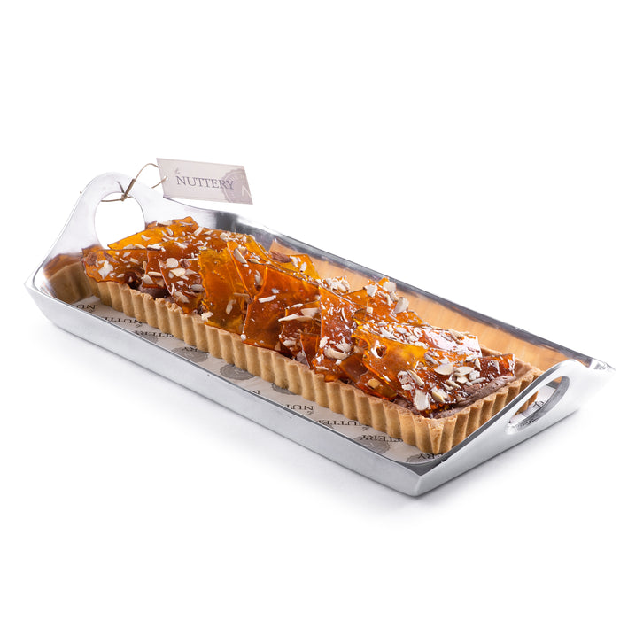 Rectangular tart topped with caramelized almond brittle, presented in a silver acrylic serving tray and tied with a branded cream ribbon, ideal for gifting or dessert tables.