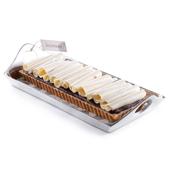 Rectangular tart layered with white chocolate curls and creamy frosting, presented in a silver acrylic serving tray and tied with a branded cream ribbon, perfect for celebrations.