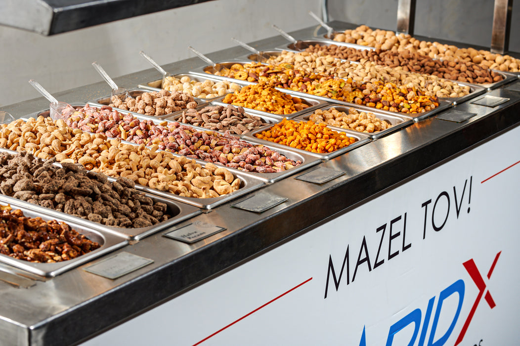 A stainless steel warming nut cart from The Nuttery, customized with a Mazel Tov and RapidX branding, featuring an assortment of warm nuts.