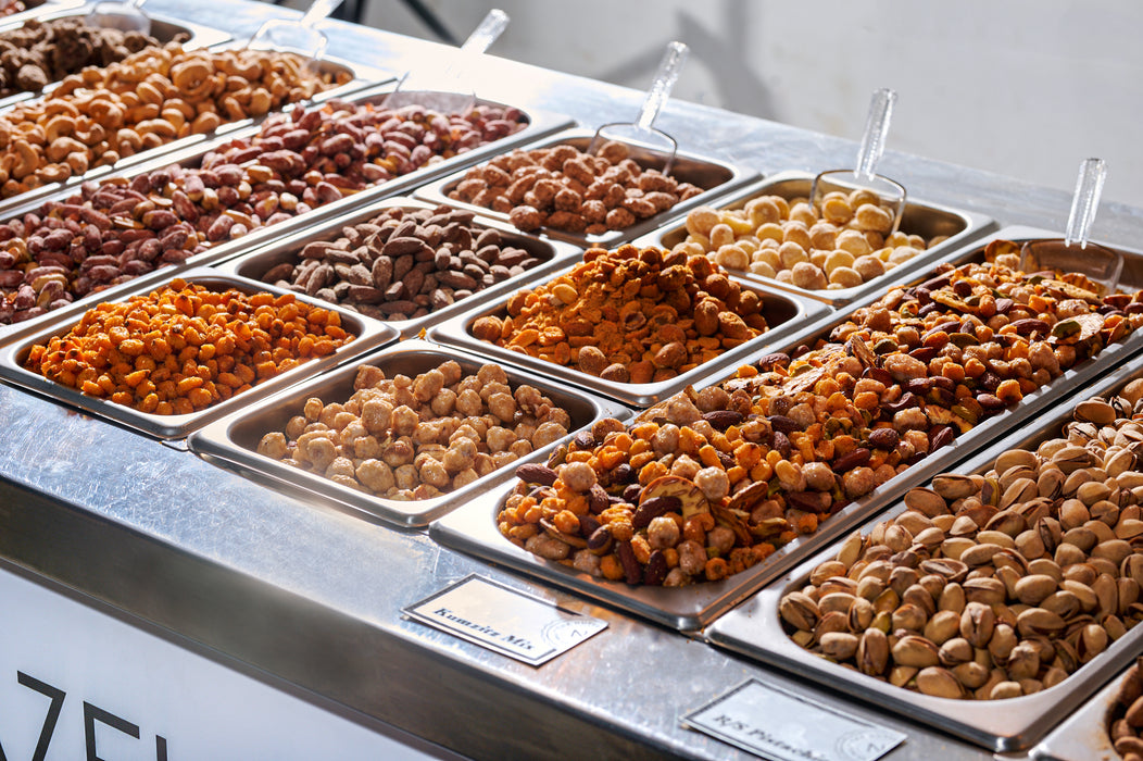 A close-up of The Nuttery’s warming nut cart, displaying a variety of roasted nuts and flavored nut mixes in stainless steel trays.