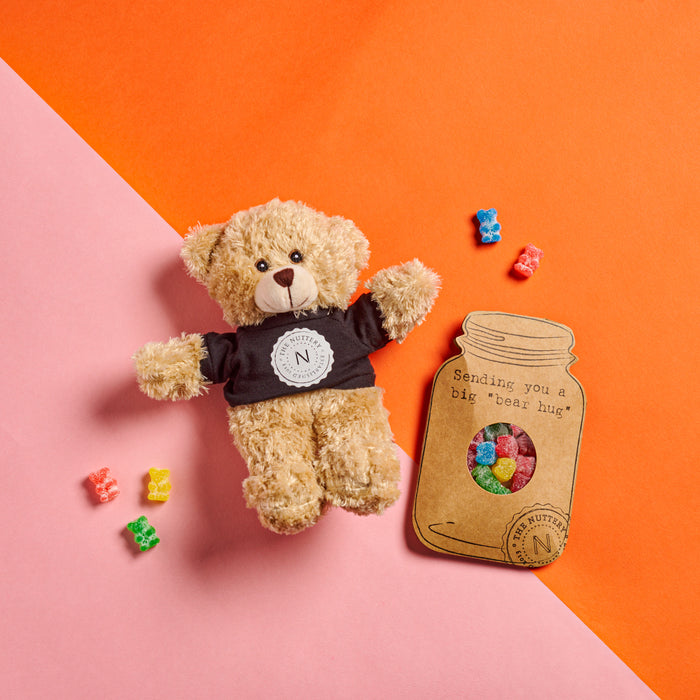 A plush teddy bear wearing a branded shirt, accompanied by a "bear hug" gift card with colorful gummy bears, making for a heartwarming and sweet present.