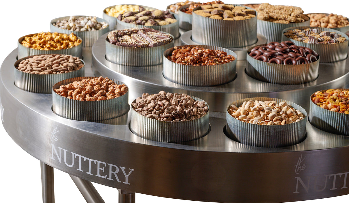 A side-angle view of The Nuttery’s round table filled with an assortment of gourmet nuts, chocolates, and candies in metal tins, designed for upscale gatherings or gifting