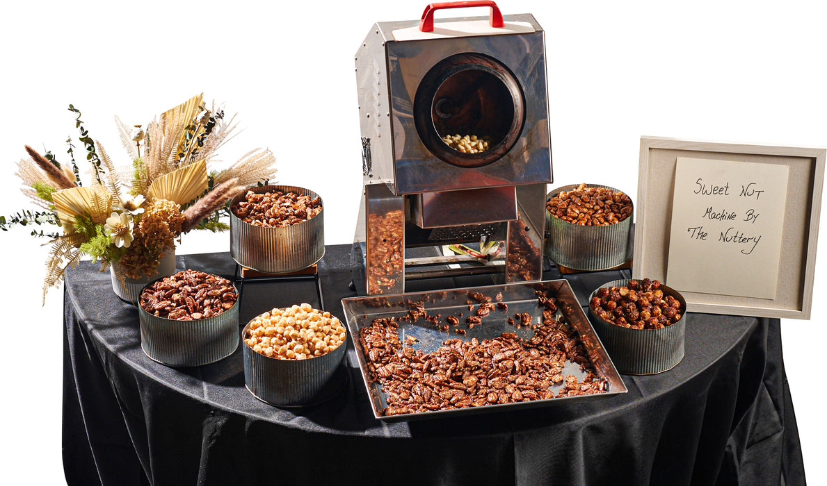 A stainless steel nut-roasting machine at The Nuttery, surrounded by trays of freshly roasted nuts, offering a gourmet experience for nut lovers.
