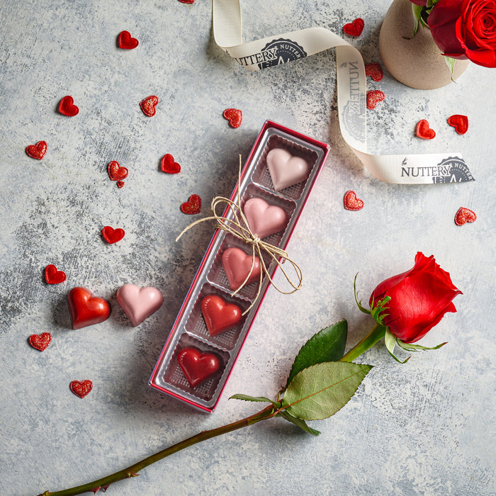 A beautifully arranged Valentine's Day truffle set featuring heart-shaped chocolates in shades of red and pink. The chocolates are nestled in a long, narrow gift box tied with twine, with scattered heart-shaped candies and a red rose adding to the romantic presentation.