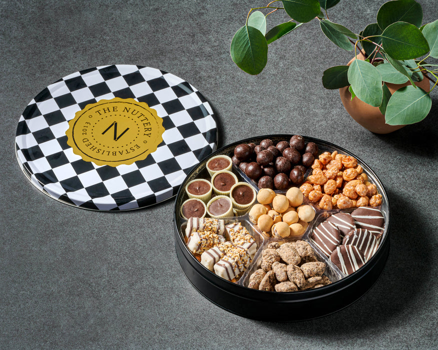 Black and white checkered tin with The Nuttery gold logo, surrounded by small potted plants, perfect for gifting or sharing gourmet treats.